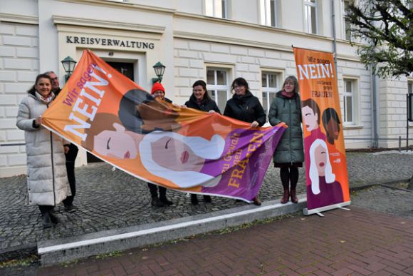 Die Fahne wird fürs Hissen vorbereitet. Die Gleichstellungsbeauftragte des Landkreises Prignitz, Christina Rätke (l.), und ihr Pendant in der Stadt Perleberg, Regine Tausendfreund (2.v.r.), hatten sich dafür vor der Kreisverwaltung verabredet. Foto: Bernd Atzenroth / Landkreis Prignitz
